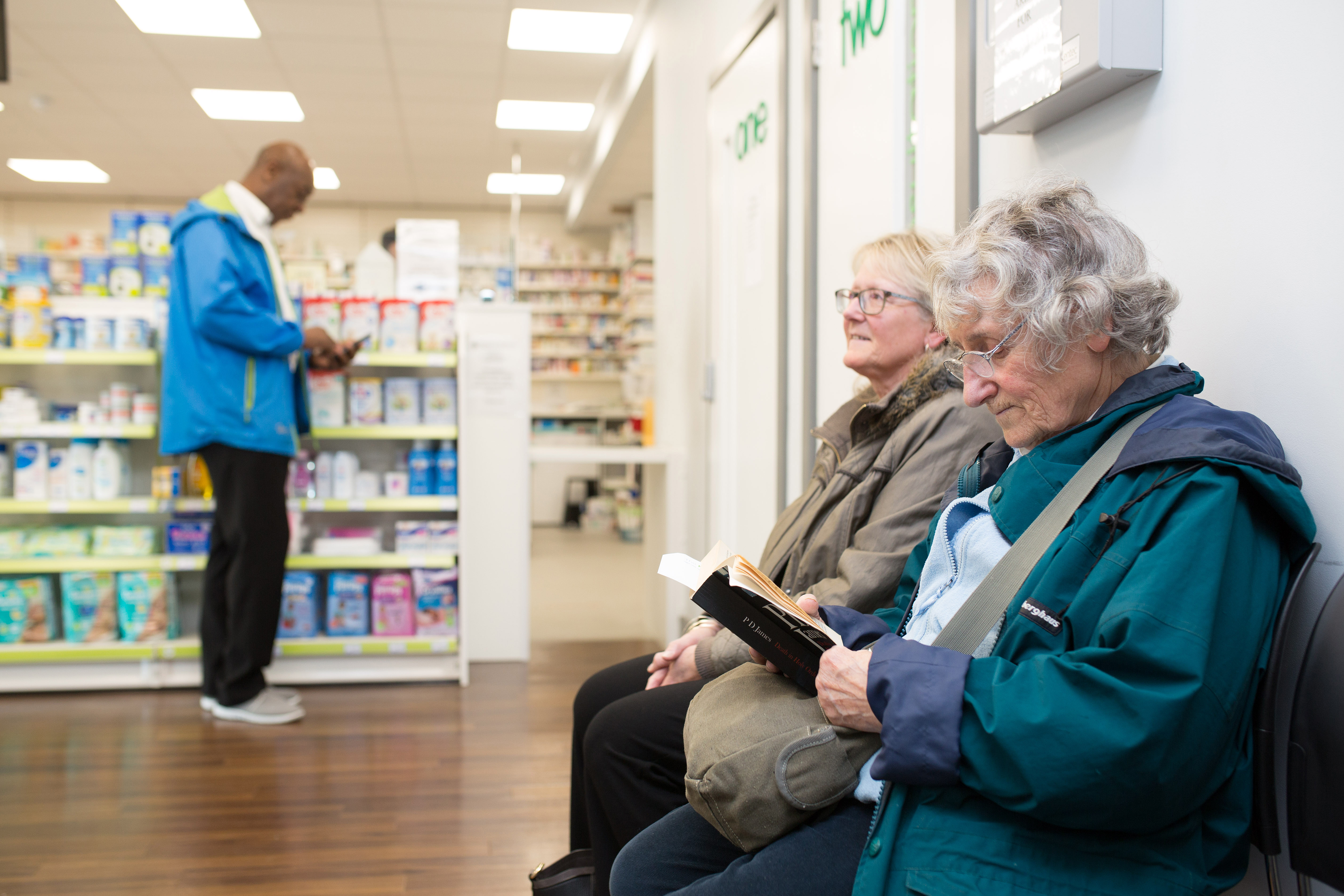 Two people sat waiting in a pharmacy while another stands up at the counter. 