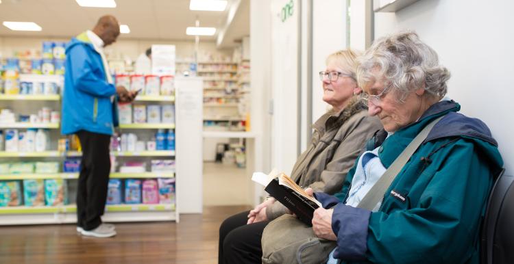 Two people sat waiting in a pharmacy while another stands up at the counter.