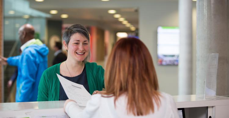 A woman smiling while talking to a receptionist holding in a document in a clinical setting.