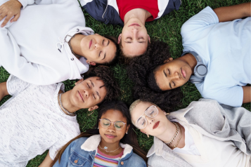 Six young people lying on grass in a circle with their heads touching.