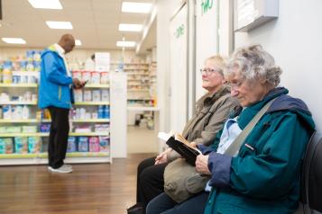 Two people sat waiting in a pharmacy while another stands up at the counter. 