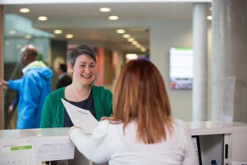 A woman smiling while talking to a receptionist holding in a document in a clinical setting.